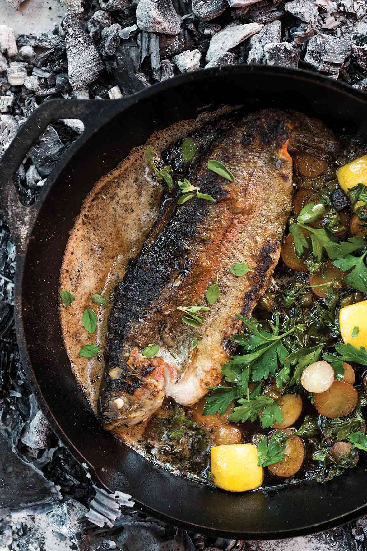 A grilled whole trout in a cast-iron skillet with sliced potatoes, parsley, and lemon