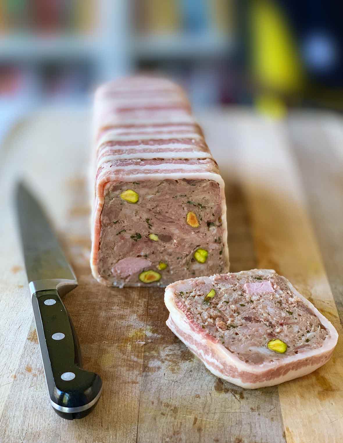 A terrine of country pate with one slice cut and resting on a wooden cutting board with a knife beside it