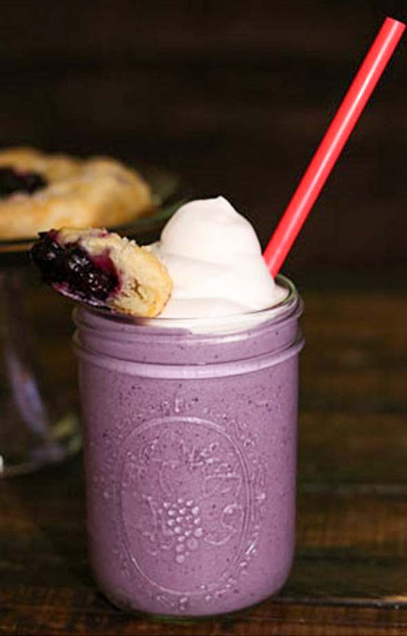 A mason jar filled with blueberry pie milkshake, topped with whipped cream and a piece of pie crust.