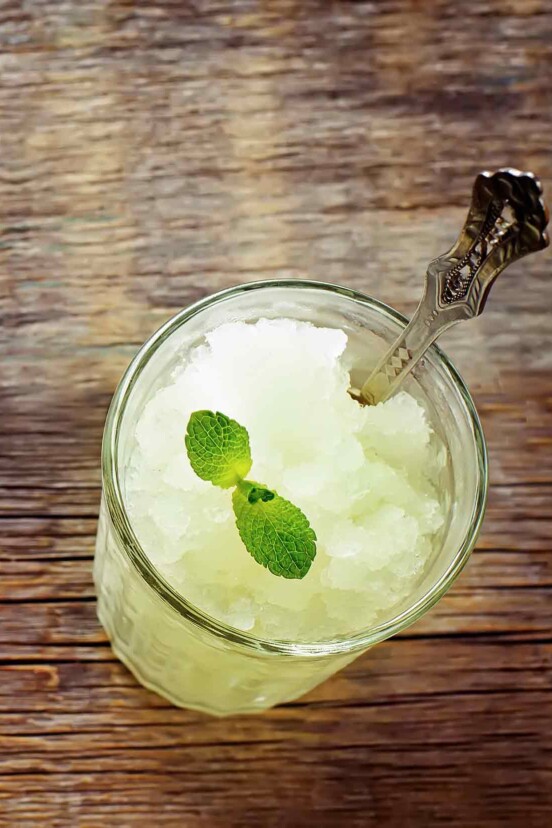 A glass filled with margarita granita, topped with a lime sprig and a spoon resting in the glass.