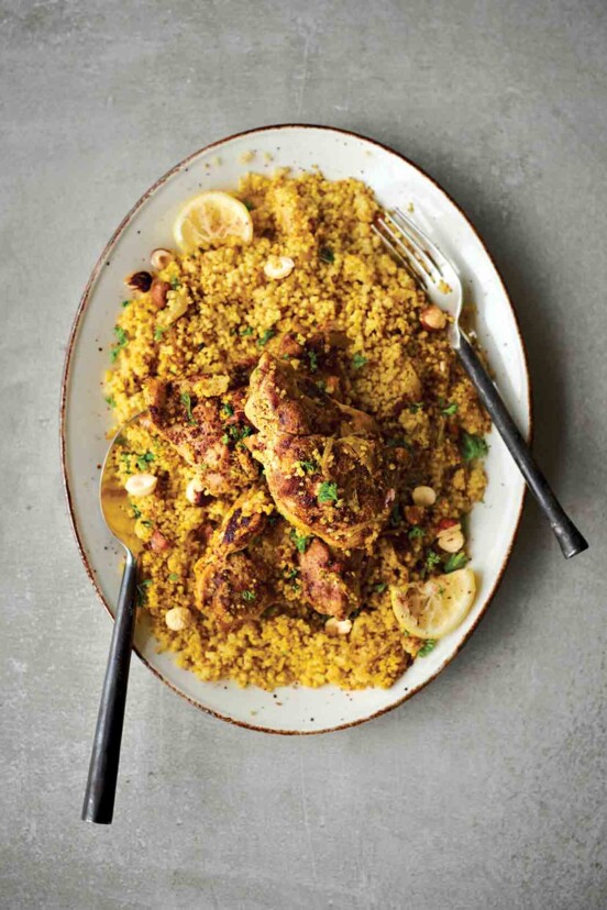 A white oval platter filled with couscous that is topped with Middle Eastern chicken thighs, lemon slices, and chopped parsley.