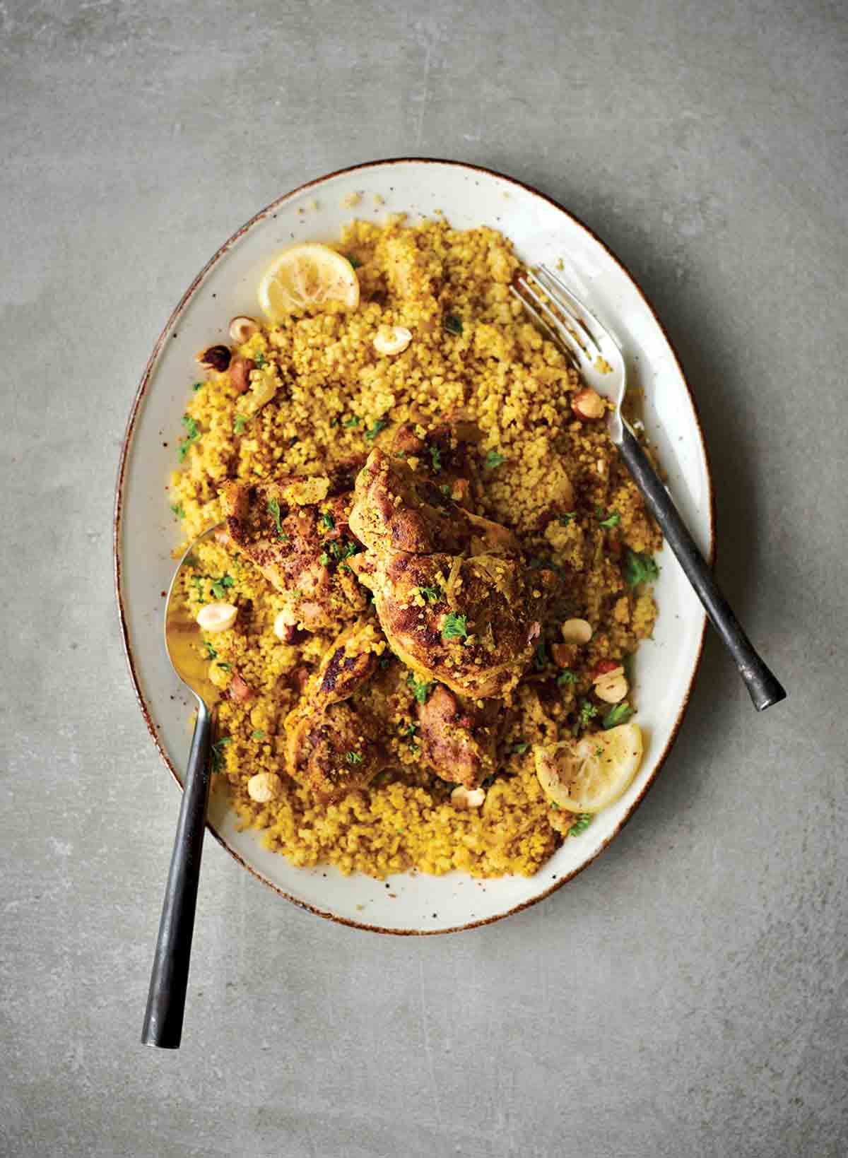 A white oval platter filled with couscous that is topped with Middle Eastern chicken thighs, lemon slices, and chopped parsley.