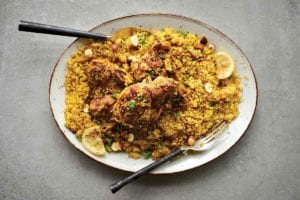 A white oval platter filled with couscous that is topped with Middle Eastern chicken thighs, lemon slices, and chopped parsley.