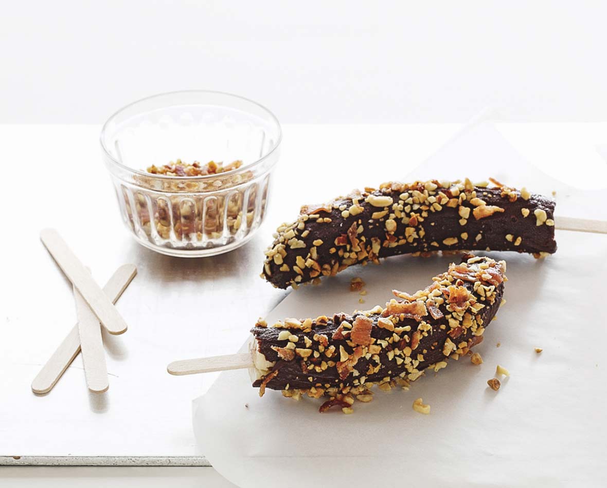 Two frozen chocolate-covered bananas on sticks with a dish of crushed peanuts and extra popsicles sticks beside them.