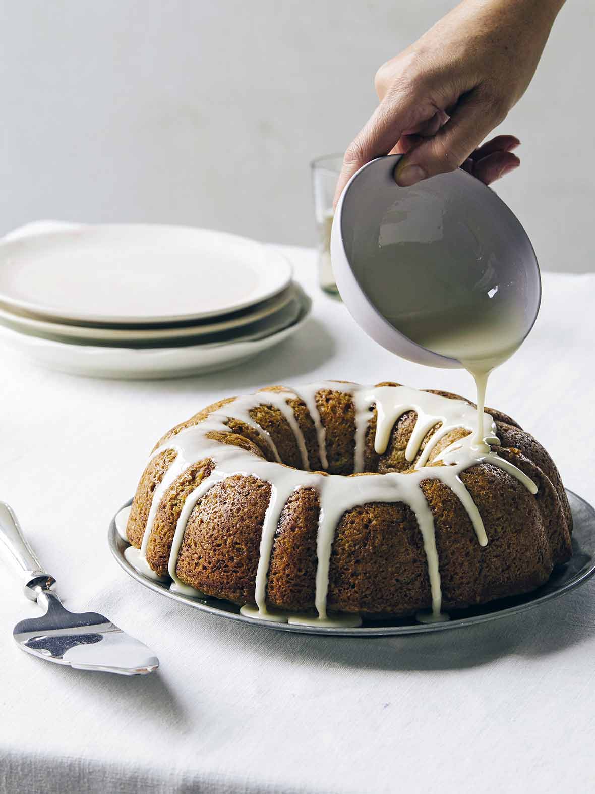 An old-fashioned zucchini cake on a silver plate with lemon glaze being drizzled over it.