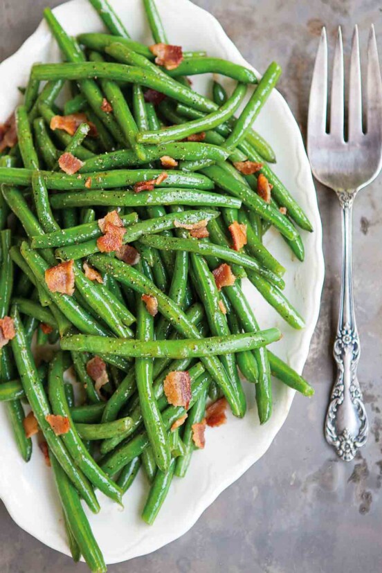 A white serving platter filled with sweet and sour green beans, topped with crumbled bacon.