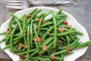 A white serving platter filled with sweet and sour green beans, topped with crumbled bacon.