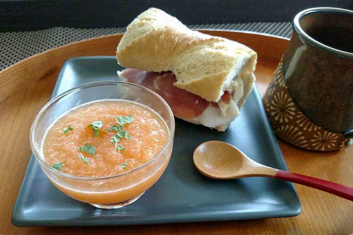 Tester's photo of cantaloupe soup with prosciutto sandwich on gray plate with wooden spoon on right.
