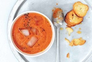 A bowl of easy gazpacho with ice cubes floating in it on a silver plate with a spoon and a few crostini.