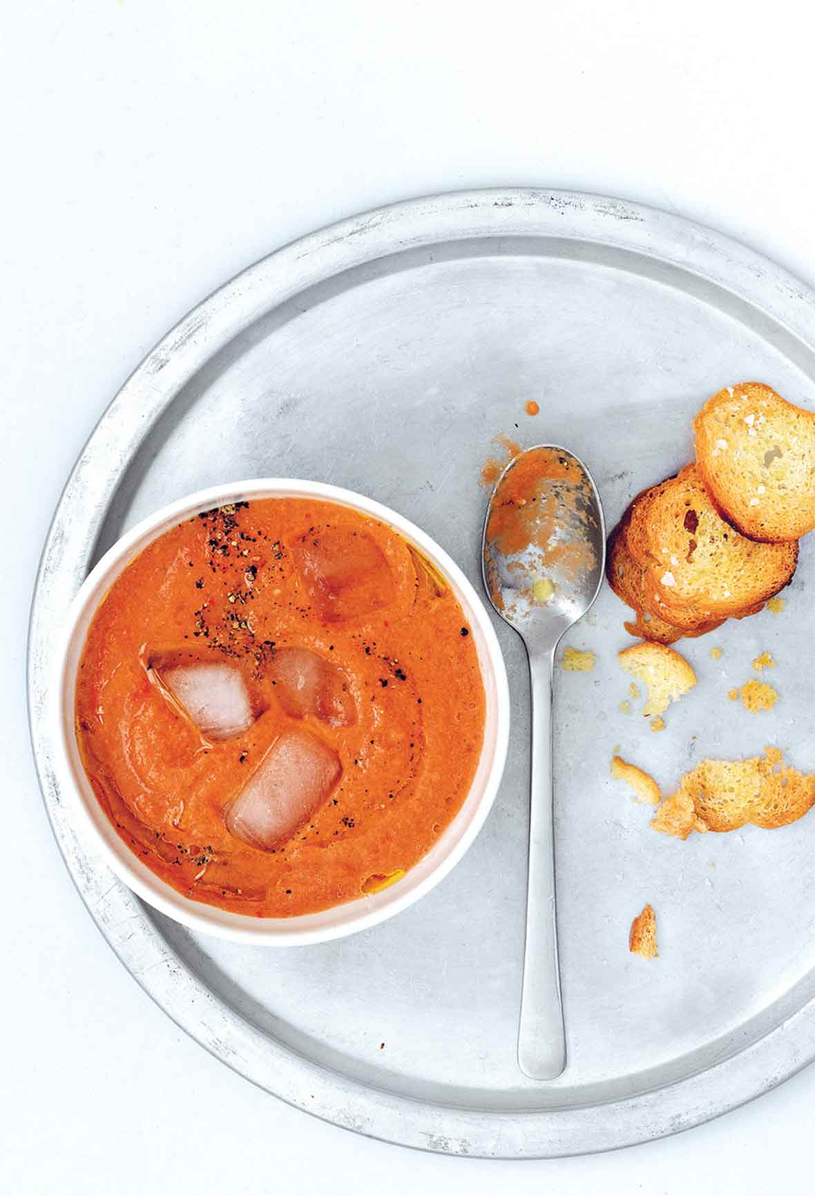 Semangkuk gazpacho sederhana dengan es batu mengambang di dalamnya, sendok di piring perak dan beberapa crostini.