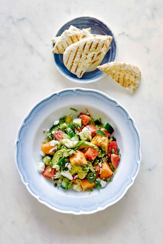 A blue and white bowl filled with melon and avocado salad with a bowl of pita beside it.