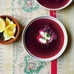 A small bowl of lemon wedges and two white bowls of deep purple summer borscht with a dollop or sour cream and sprinkling of chives in each.