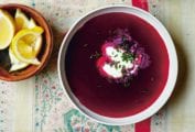 A small bowl of lemon wedges and two white bowls of deep purple summer borscht with a dollop or sour cream and sprinkling of chives in each.