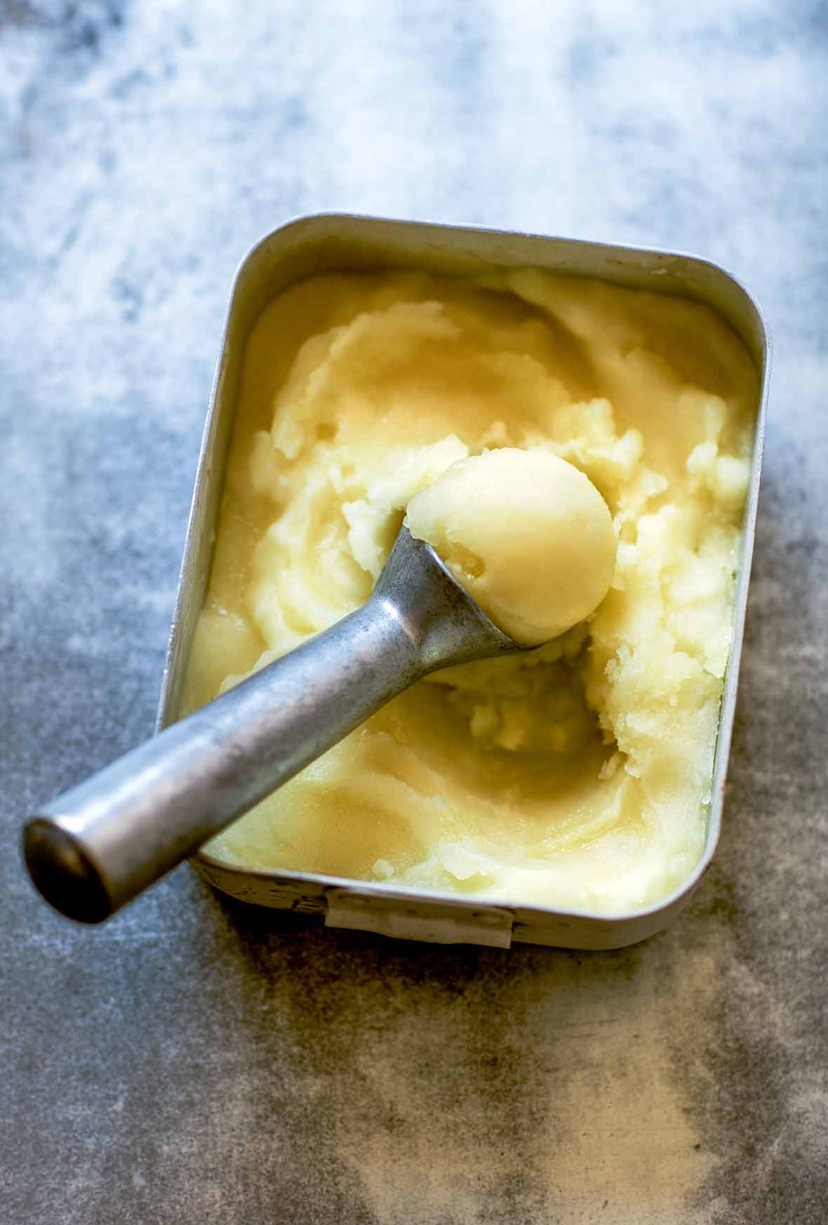 A metal container filled with tart lemon sorbet and a metal ice cream scoop resting inside.