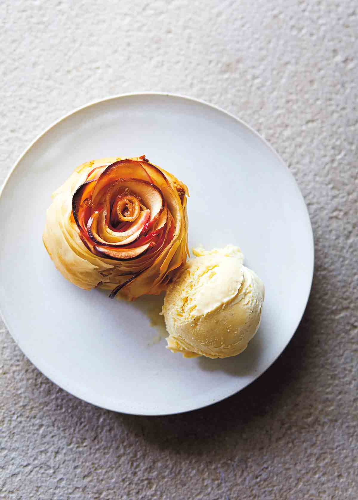 En äppelrostårta, formad för att se ut som en ros med en kula glass bredvid på en vit tallrik.