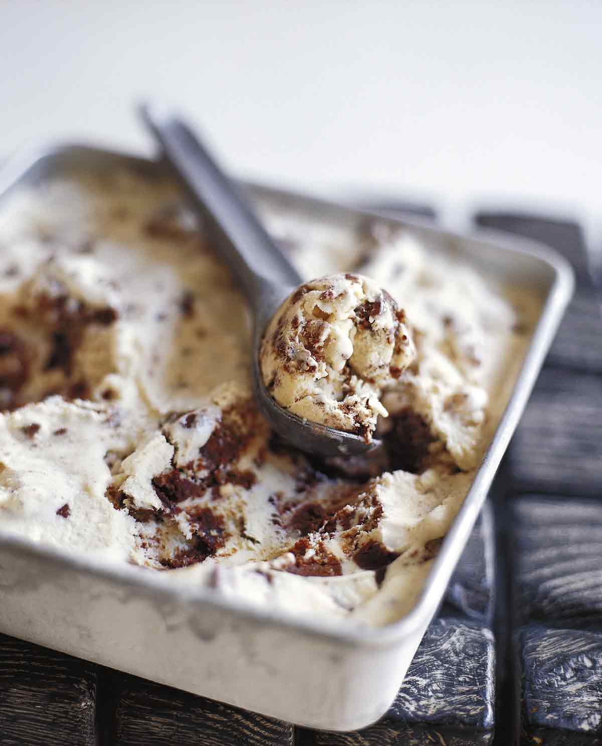 A square metal dish filled with brownie ice cream and a metal ice cream scoop removing a scoop from the dish.