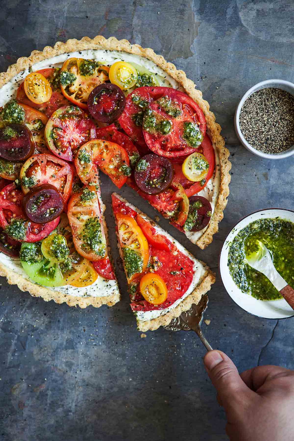 A person removing a slice of fresh tomato tart with a lifter and a bowl of pesto and black pepper on the side.