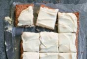 A frosted apple cake on a sheet of wax paper that has been cut into 12 squares with a knife resting beside.