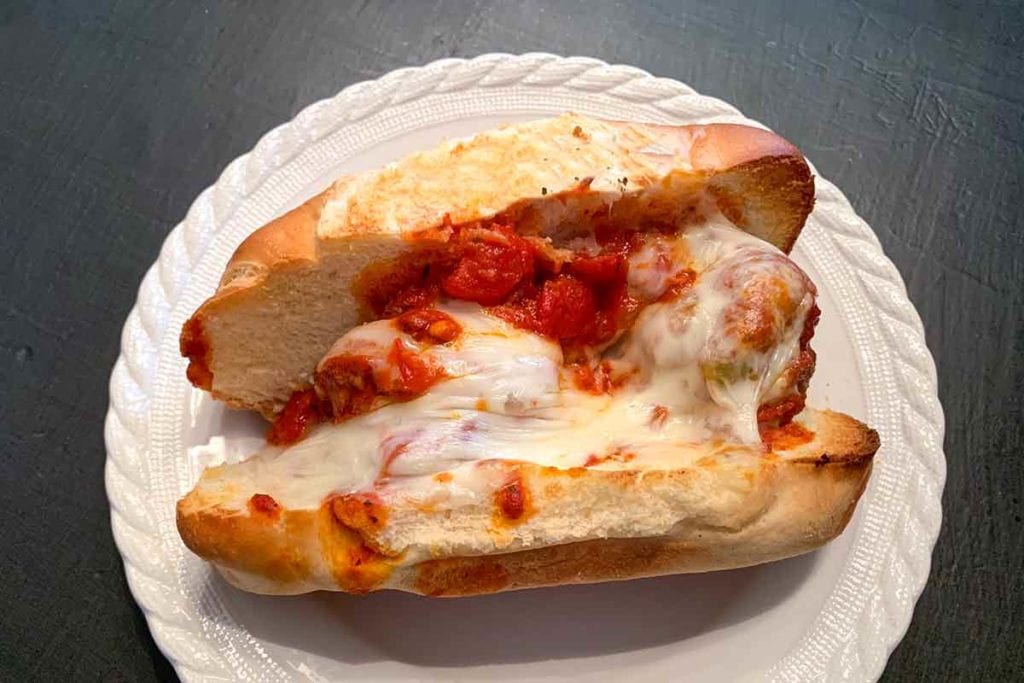A harissa meatballs sandwich, made with three meatballs stuffed in a sub roll along with cheese and topped with tomato sauce on a white plate.
