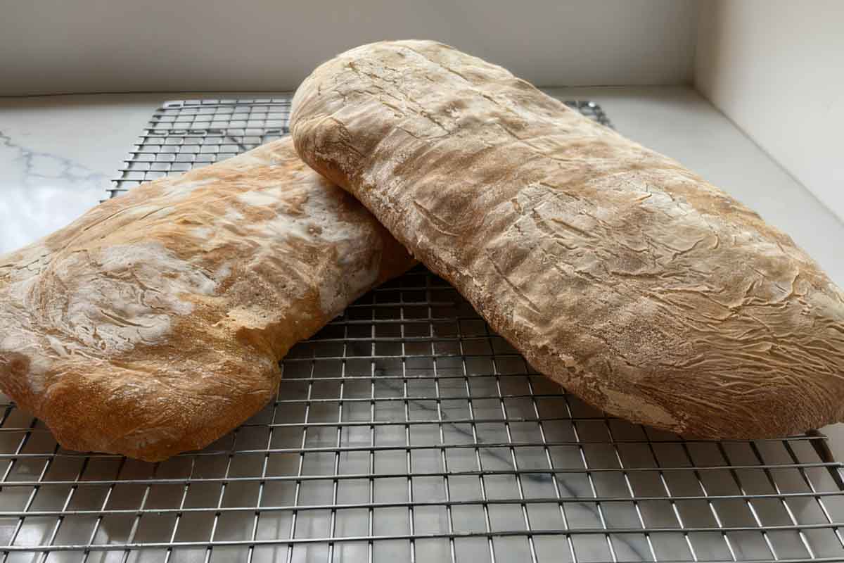 Two flowery loaves of ciabatta cooling on a wire rack by a window.