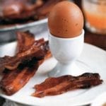 Three strips of maple candied bacon on a white plate along with a boiled egg in an egg cup.