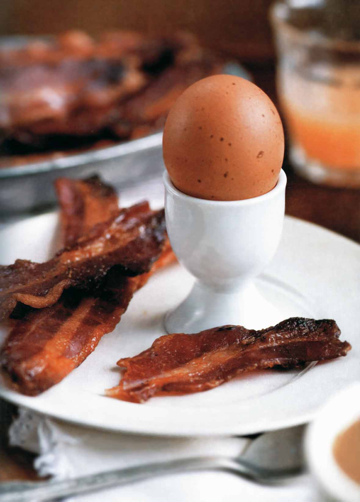Three strips of maple candied bacon on a white plate along with a boiled egg in an egg cup.