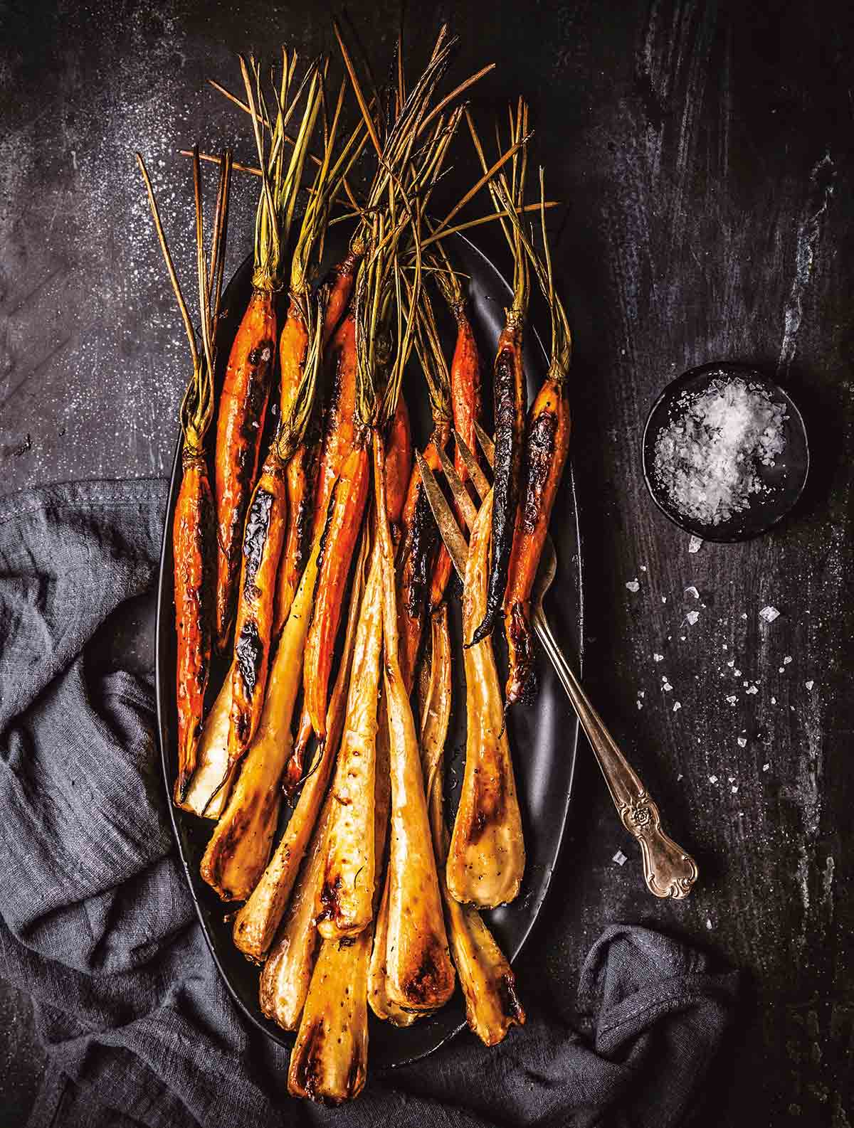 Maple glazed carrots with bourbon and parsnips on a black oval platter with a dish of salt beside it.