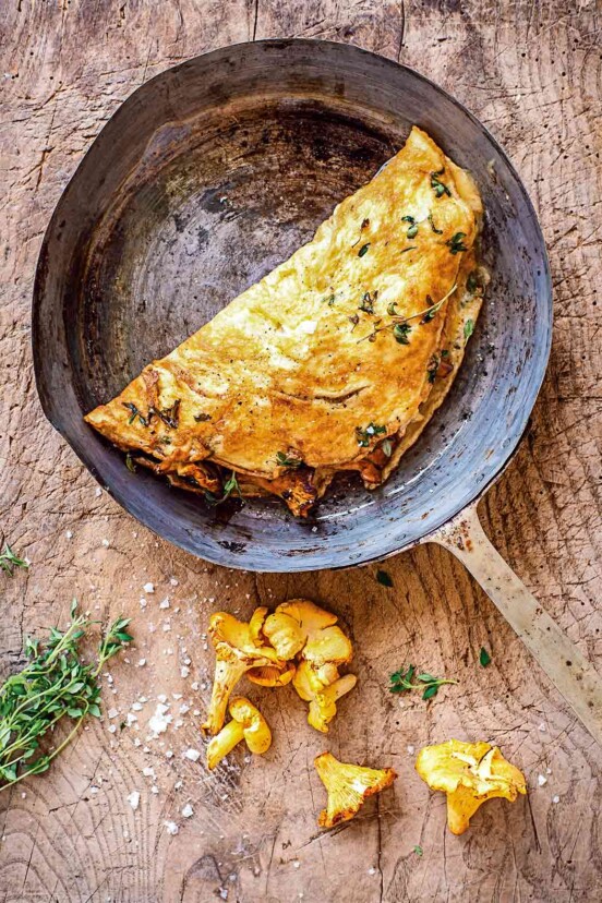 An omelet with chanterelles in a metal skillet with two chanterelle mushrooms lying beside the skillet.