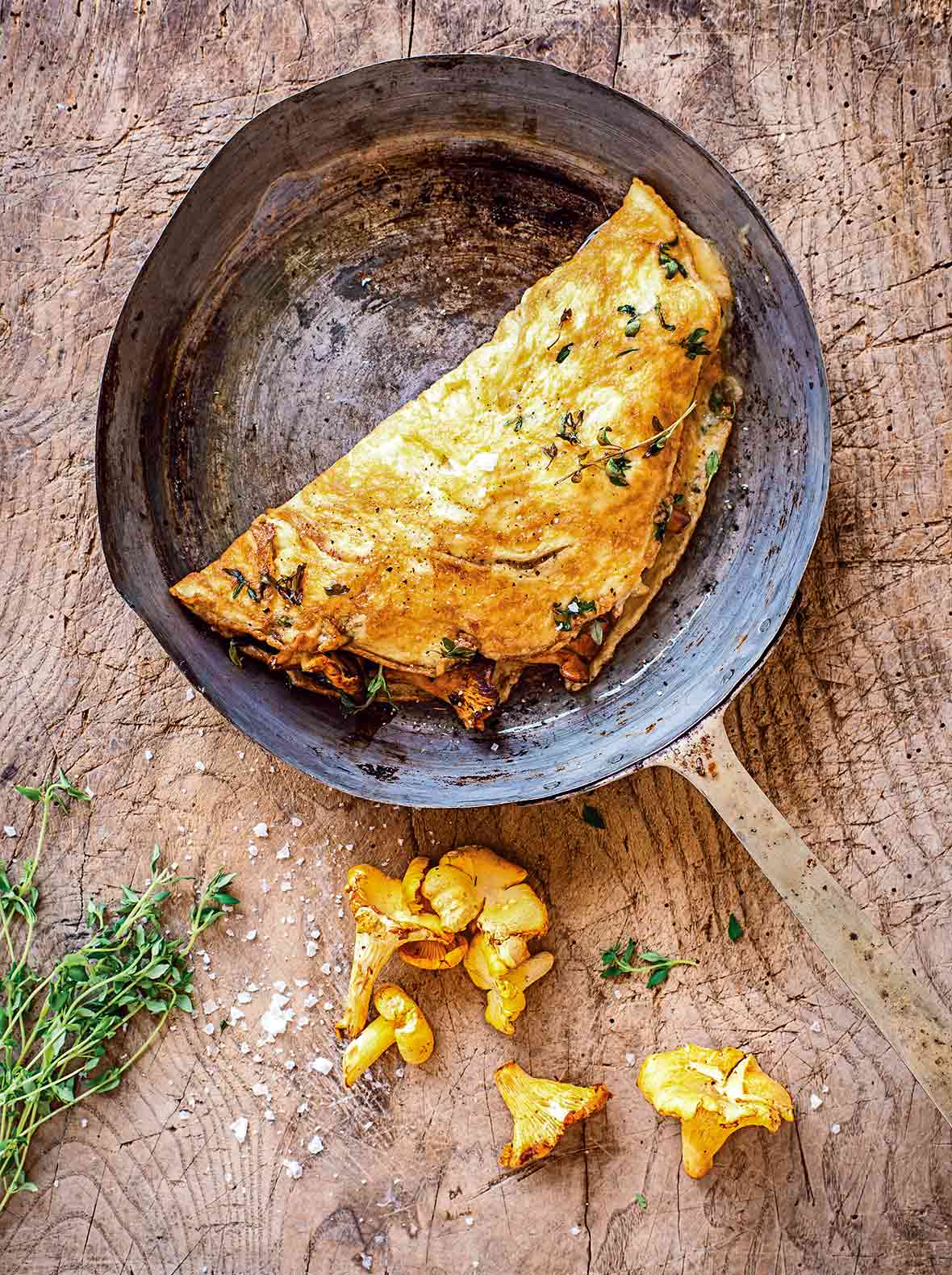 An omelet with chanterelles in a metal skillet with two chanterelle mushrooms lying beside the skillet.