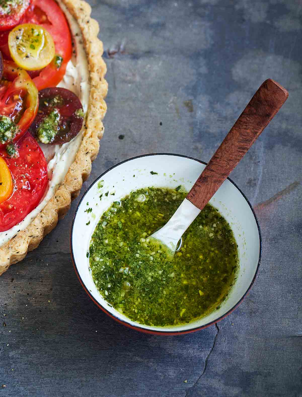 A bowl of pesto vinaigrette with a spoon inside beside a fresh tomato tart.