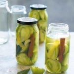 Three jars filled with pickled green tomatoes and a cinnamon stick with two empty jars in the background.