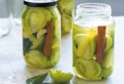 Three jars filled with pickled green tomatoes and a cinnamon stick with two empty jars in the background.