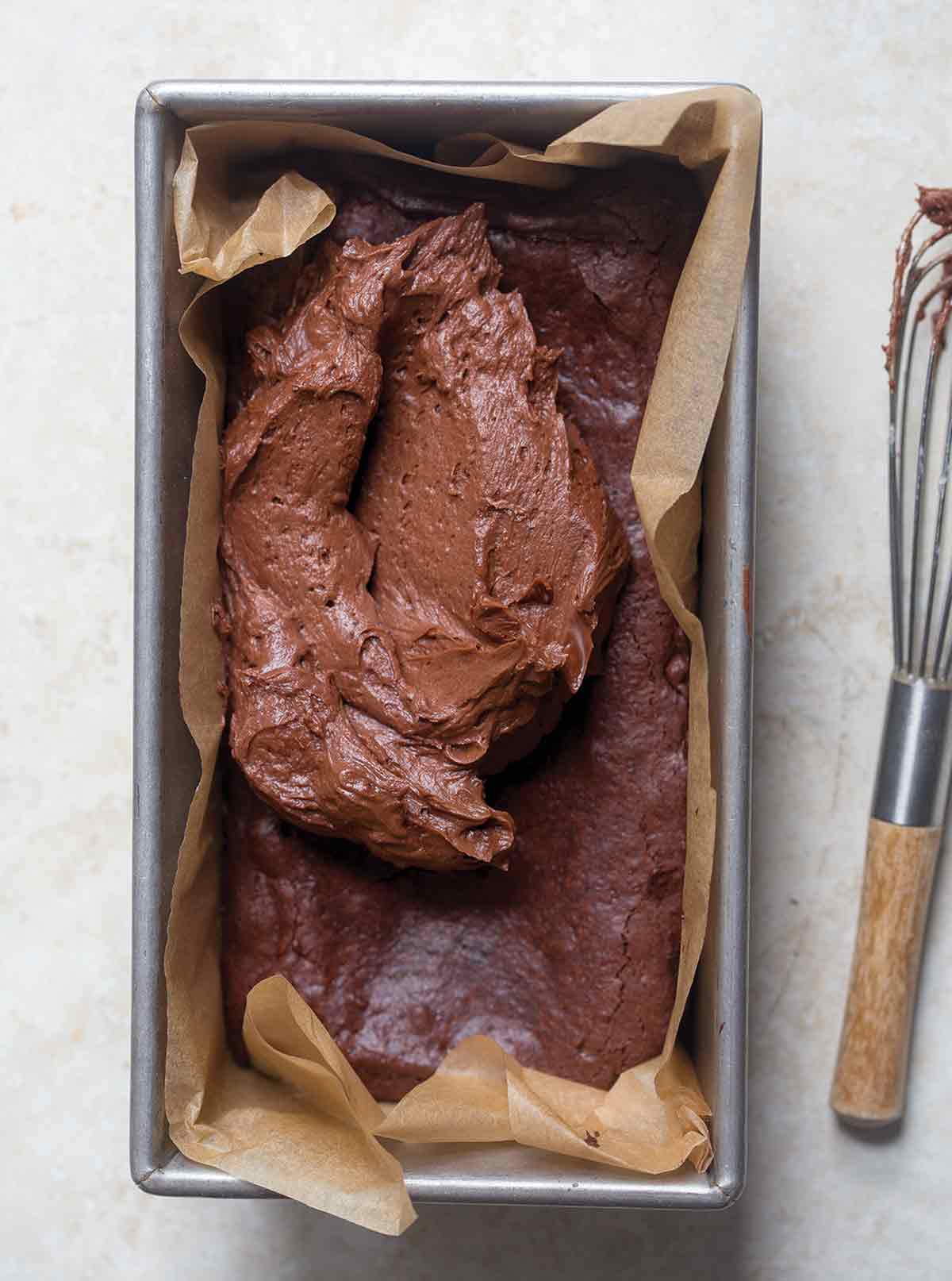 A parchment-lined loaf pan filled with partially frosted brownies in small portions and a whisk on the side.