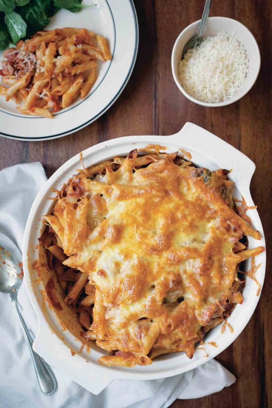 A white round baking dish filled with baked ziti and topped with melted cheese, and a dish of Parmesan on the side.