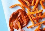 A hand dipping fries into a splatter of bomb hot sauce with a pile of fries scattered beside the sauce.