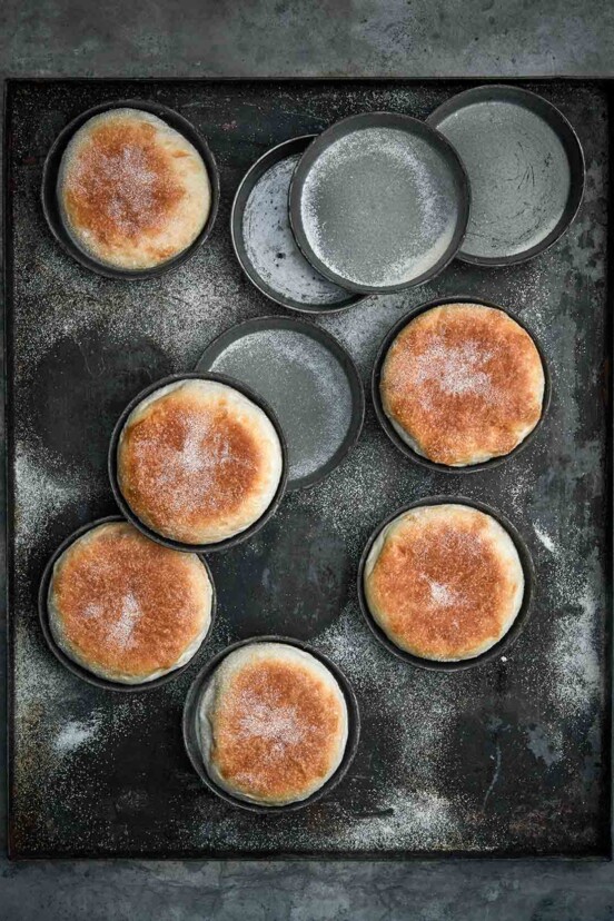 Six homemade English muffins in tins on a baking sheet and four empty tins.