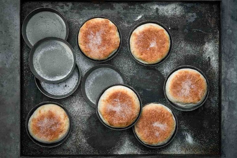 Six homemade English muffins in tins on a baking sheet and four empty tins.
