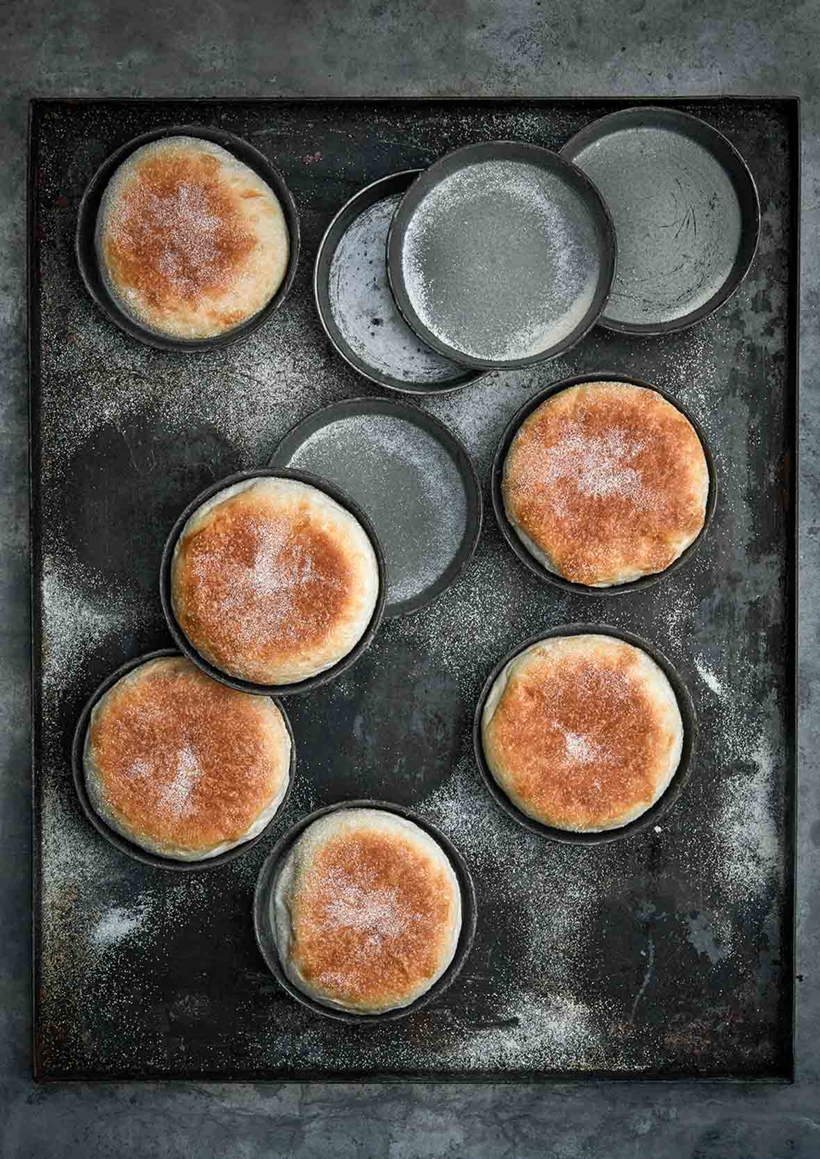 Six homemade English muffins in tins on a baking sheet and four empty tins.