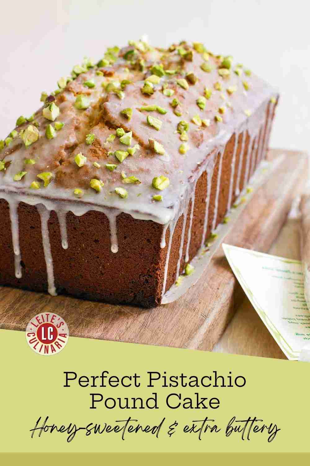 A tall golden pound cake on a wooden board with white glaze dripping down and crushed green pistachios on top.