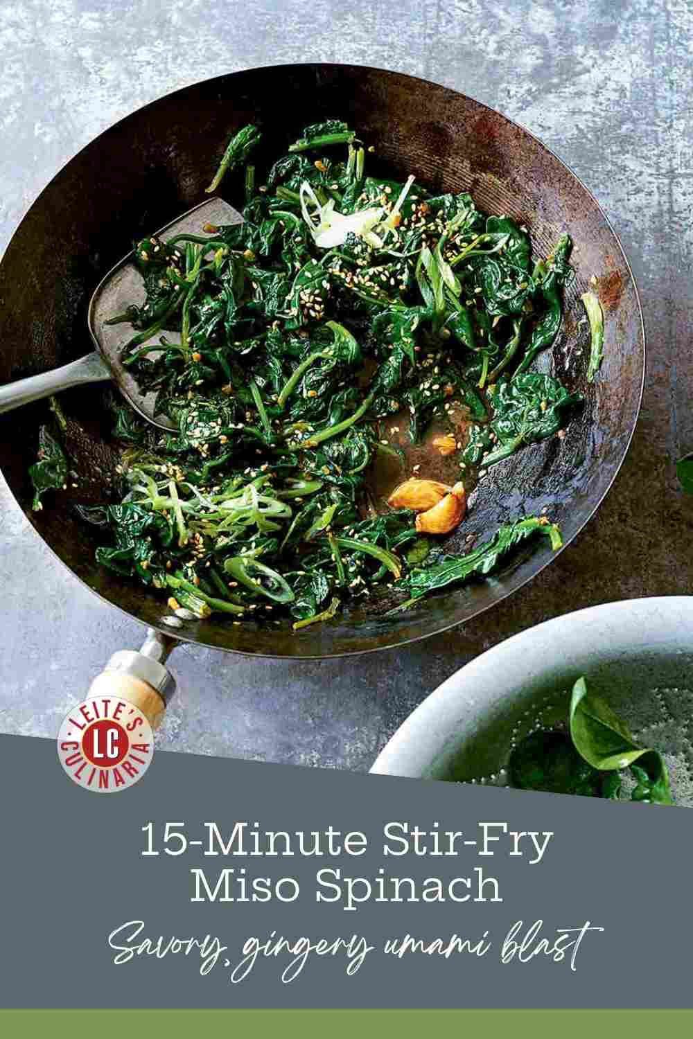 Sautéed baby spinach wilting in a dark wok with garlic cloves and toasted sesame seeds on a gray stone surface.