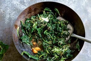 A wok filled with stir-fried spinach with miso and ginger and a colander with fresh spinach beside the wok.