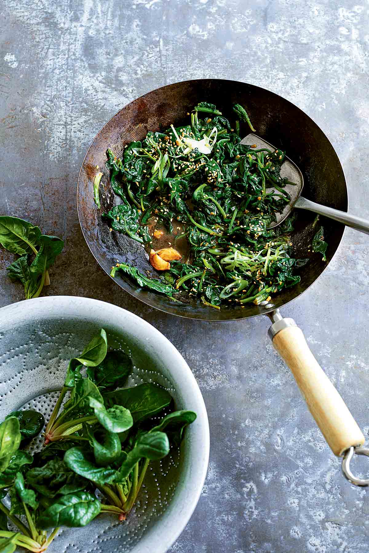 A wok filled with stir-fried spinach with miso and ginger and a colander with fresh spinach beside the wok.