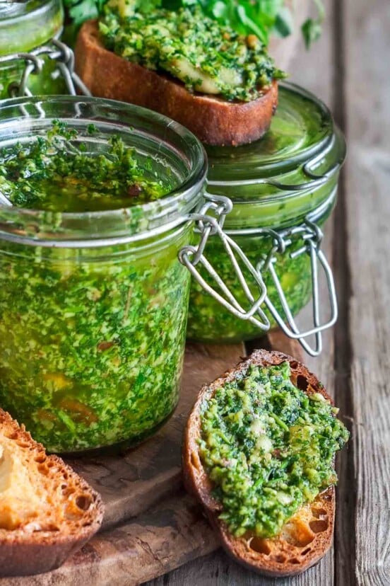 Several jars of cilantro sauce and a few toasted bread rounds topped with the cilantro sauce on a wooden board.