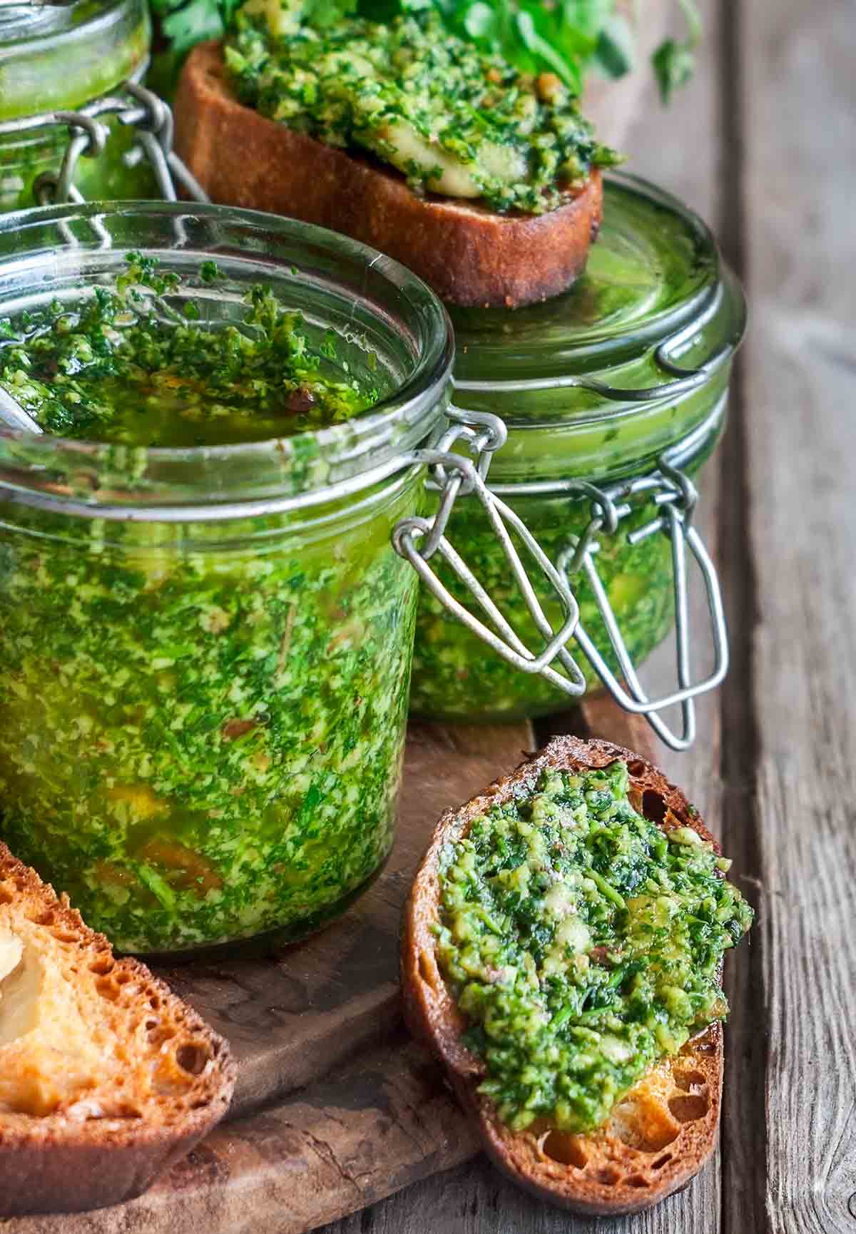 Several jars of cilantro sauce and a few toasted bread rounds topped with the cilantro sauce on a wooden board.
