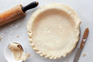 A partially crimped perfect flaky pie crust in a pie plate with a knife, rolling pin, and bowl of crust scraps beside it.
