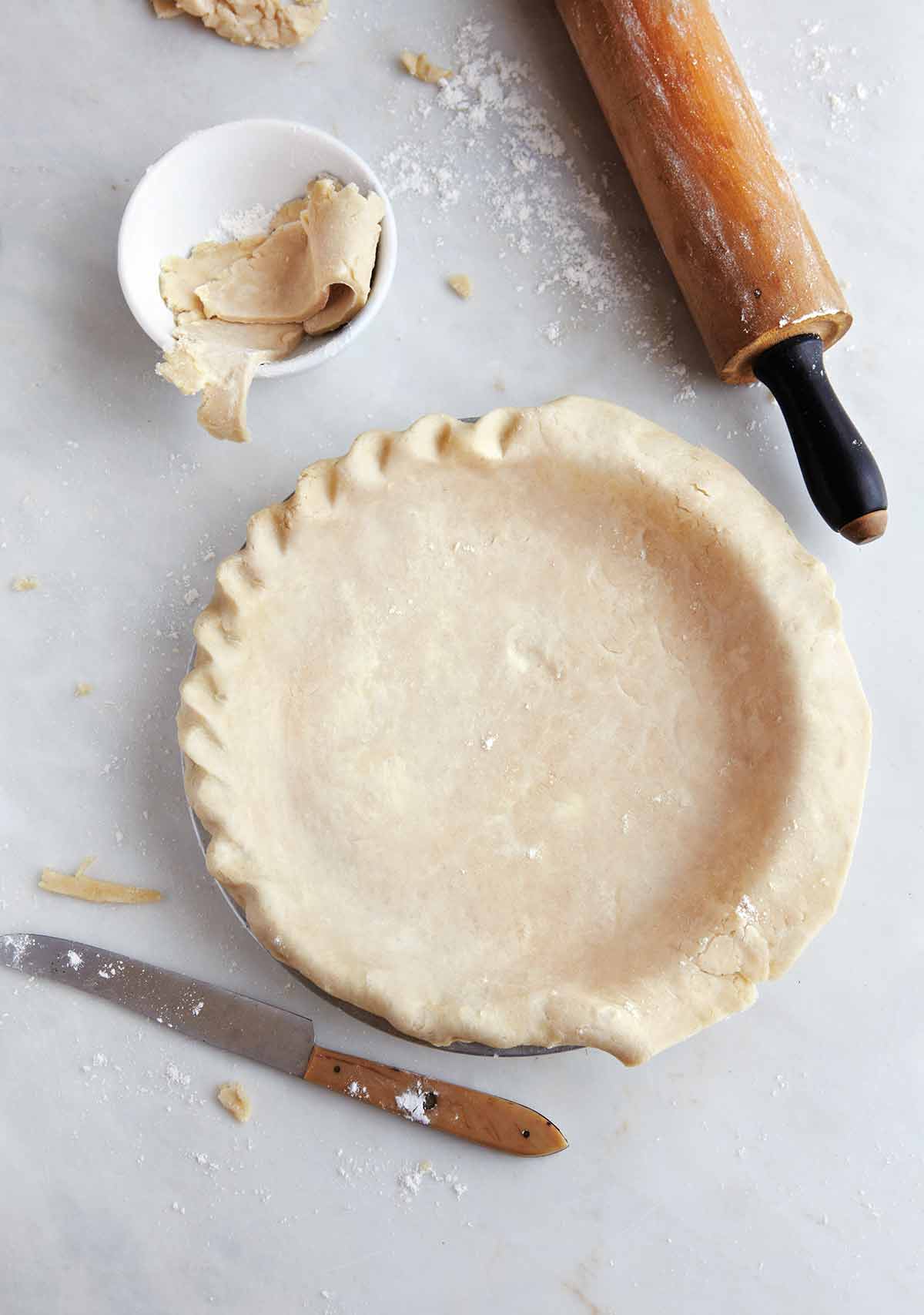A partially crimped perfect flaky pie crust in a pie plate with a knife, rolling pin, and bowl of crust scraps beside it.