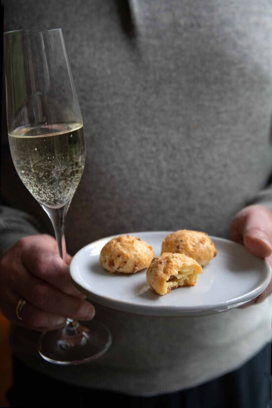 A man holding a glass of Prosecco and a plate with three gougeres.