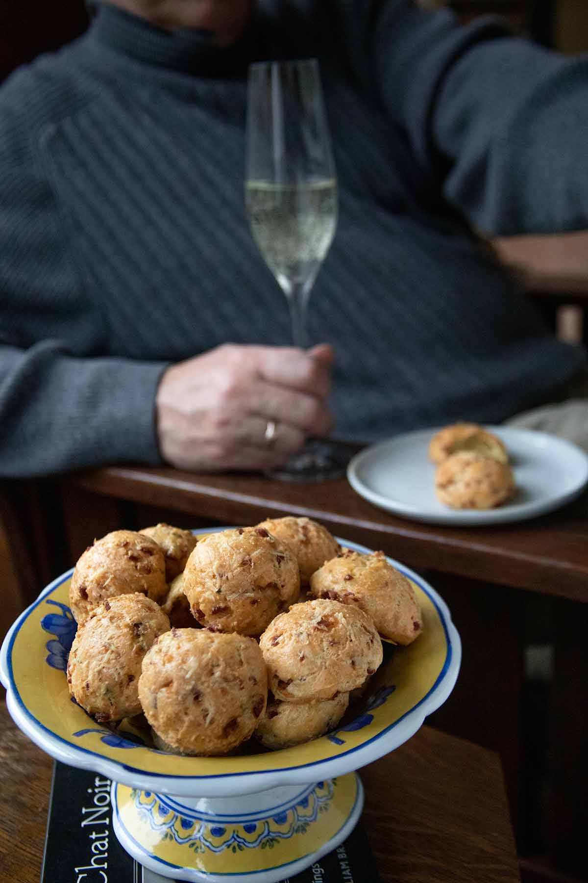A man holding a glass of Prosecco with a plate of two gougeres and a dish filled with many gougeres beside him.
