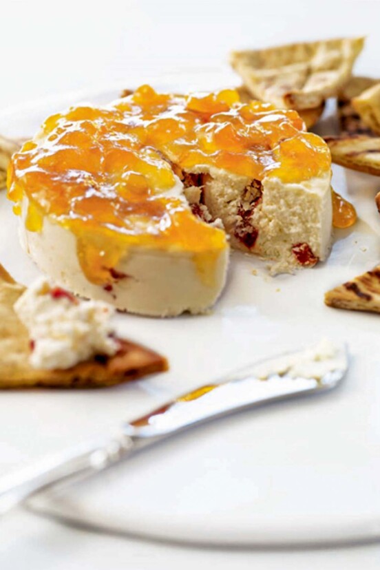 A round of red pepper cheesecake, topped with apricot jam and surrounded by crackers.