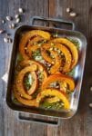 A metal roasting pan filled with roast squash, feta, and pistachios on a wooden table with pistachios scattered around it.